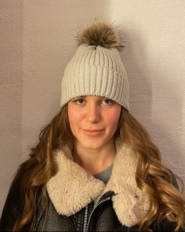 Beige ribbstickad mössa med pälstofs, sedd på modell.
